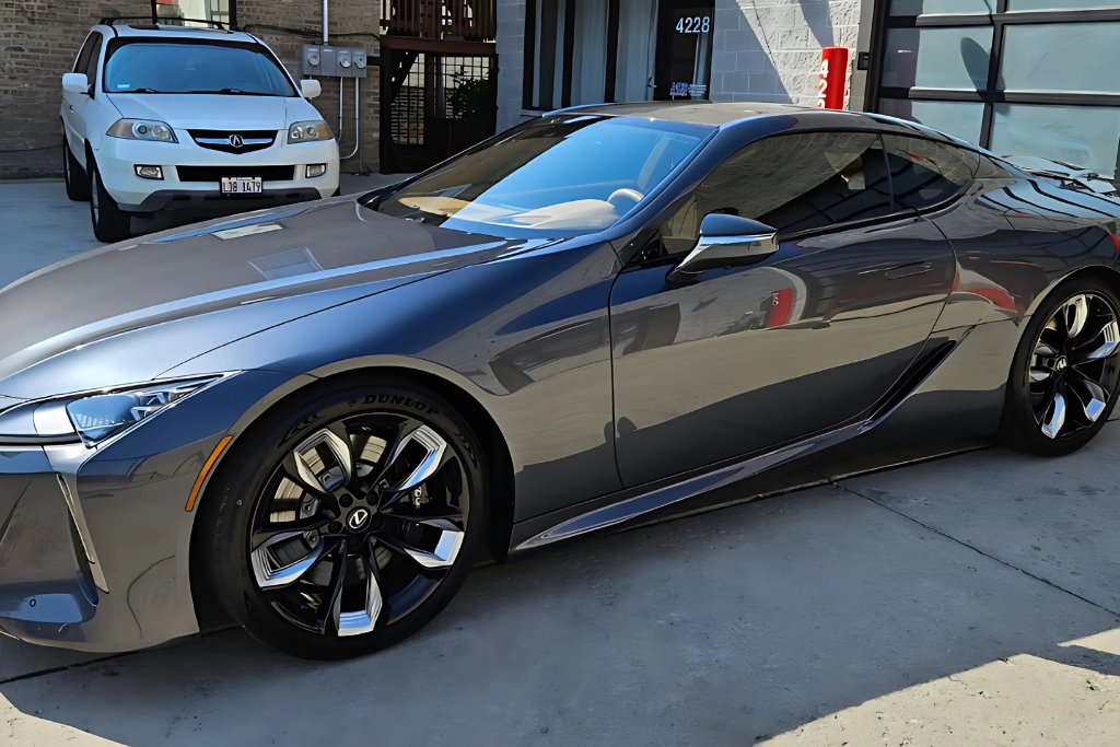 A sleek, dark gray sports car with a glossy, reflective finish sits in an outdoor setting, showcasing the effects of ceramic coating maintenance. The car's smooth, polished surface reflects its surroundings, highlighting the coating's ability to repel dirt and maintain shine.