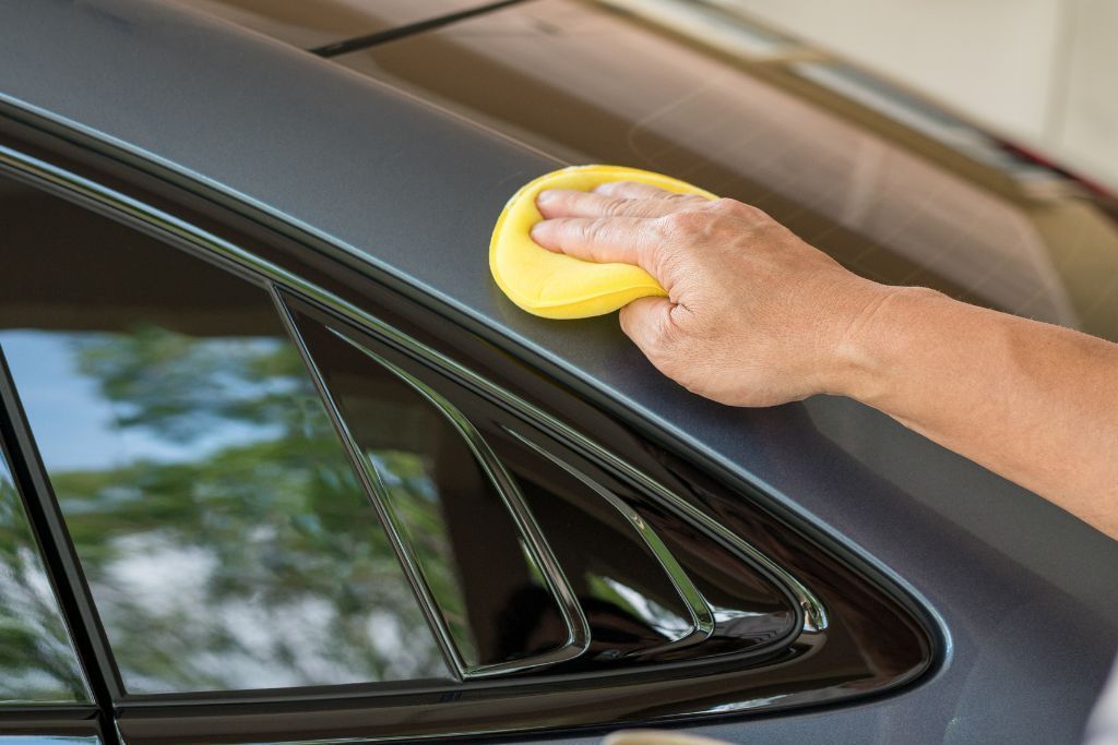 A person uses a yellow applicator pad to apply ceramic coating to the side of a car, ensuring even coverage over the vehicle's surface.