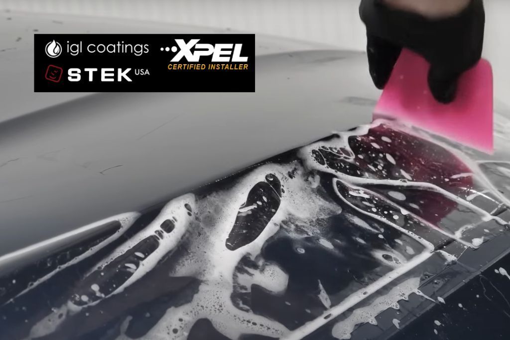 A close-up of a car surface being treated with a protective film. A person wearing a glove uses a pink squeegee to smooth out the film over soapy water. Logos for IGL Coatings, STEK USA, and XPEL Certified Installer are visible in the upper left corner, indicating the brands involved in the process.