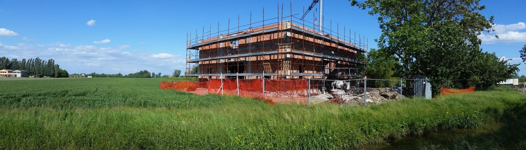 Un edificio in mattoni in costruzione in un campo in una giornata di sole con cielo azzurro. Sono presenti impalcature e reti arancioni.