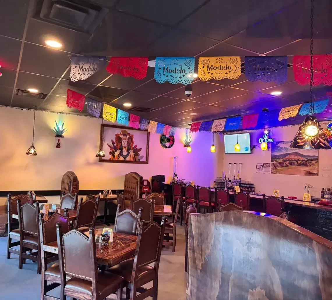 A restaurant dining area featuring wooden chairs and tables, decorated with colorful papel picado banners on the ceiling.