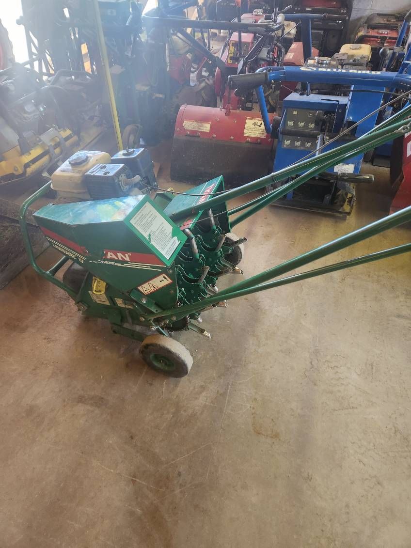 Green agricultural machine with long hoses in a workshop, surrounded by other farm equipment.