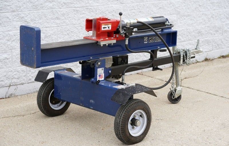 Blue log splitter on wheels with red engine, parked on a concrete driveway against a wall.