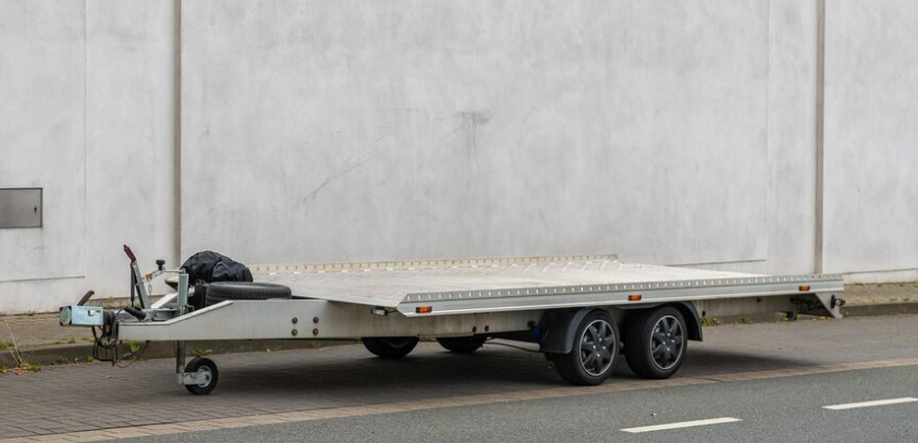 Flatbed trailer with a beige platform parked beside a concrete wall on a street.