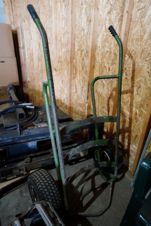 Two-wheeled hand truck with green handles leaning against a wooden wall in a cluttered storage area