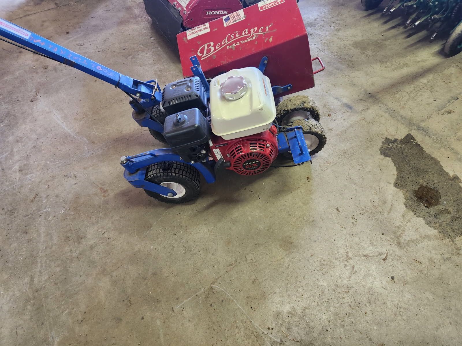 Blue walk-behind tiller with red engine on a concrete floor next to a red box.