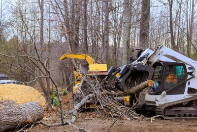 Yellow excavator and white skid steer clearing rubble in a wooded construction site