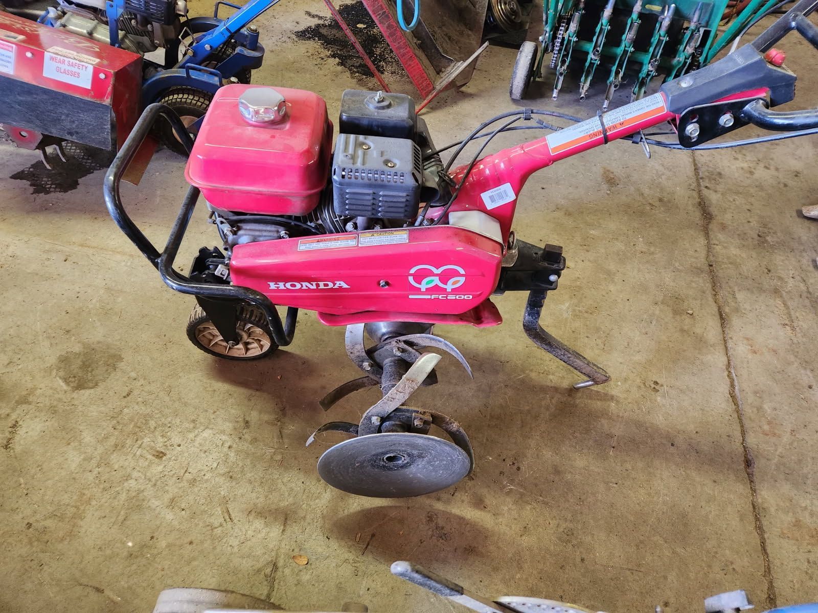 Red Honda tiller with black handlebars in a workshop, showing the engine and metal tines.