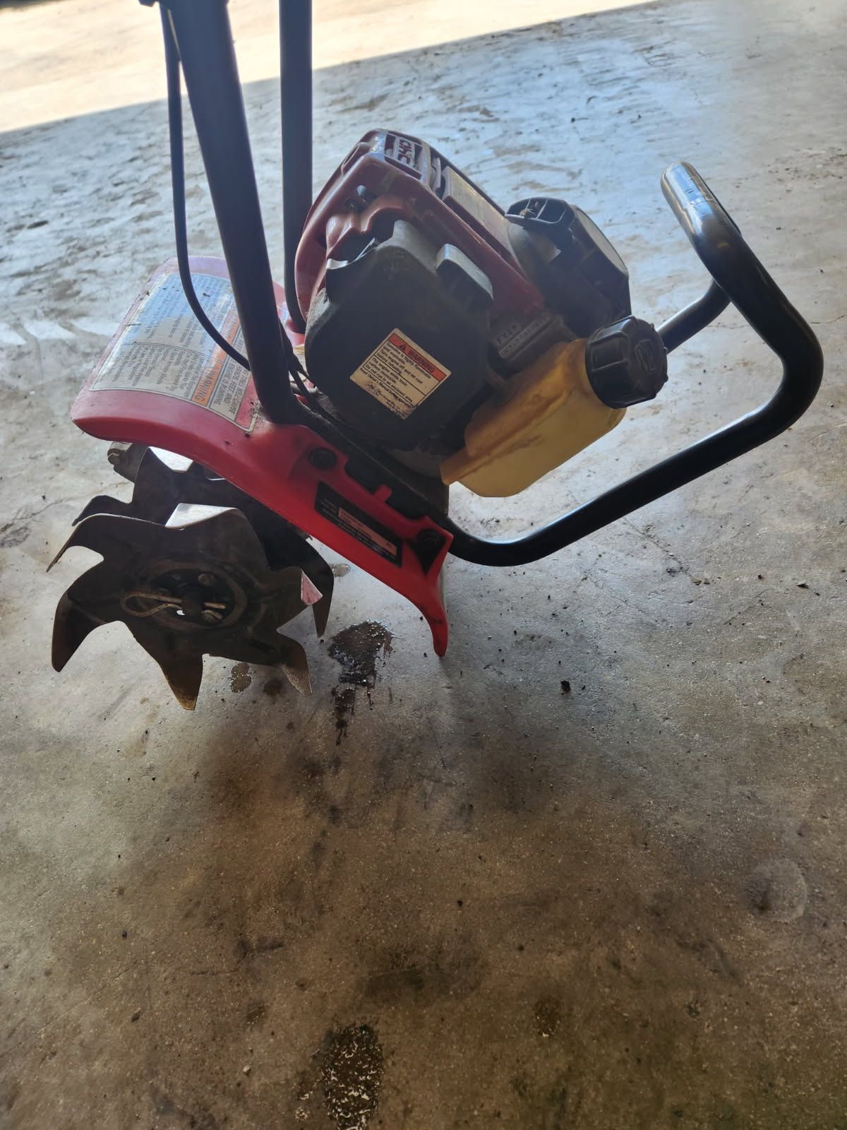 Red tiller with black handles on a dusty concrete floor, viewed from the side.