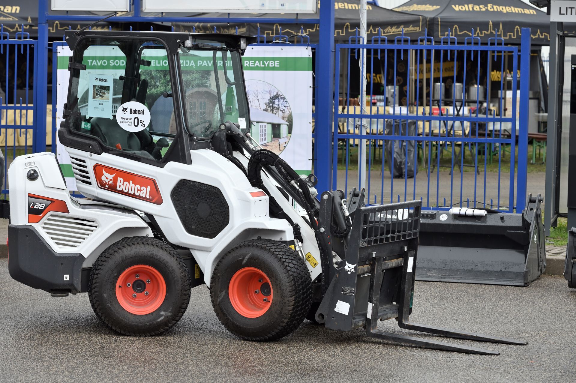 White and black Bobcat skid-steer loader with forks parked in front of a blue metal fence