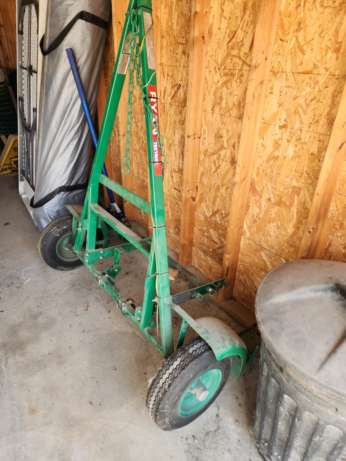 Green hand truck leaning against plywood wall in a garage beside a trash can and folded cart.