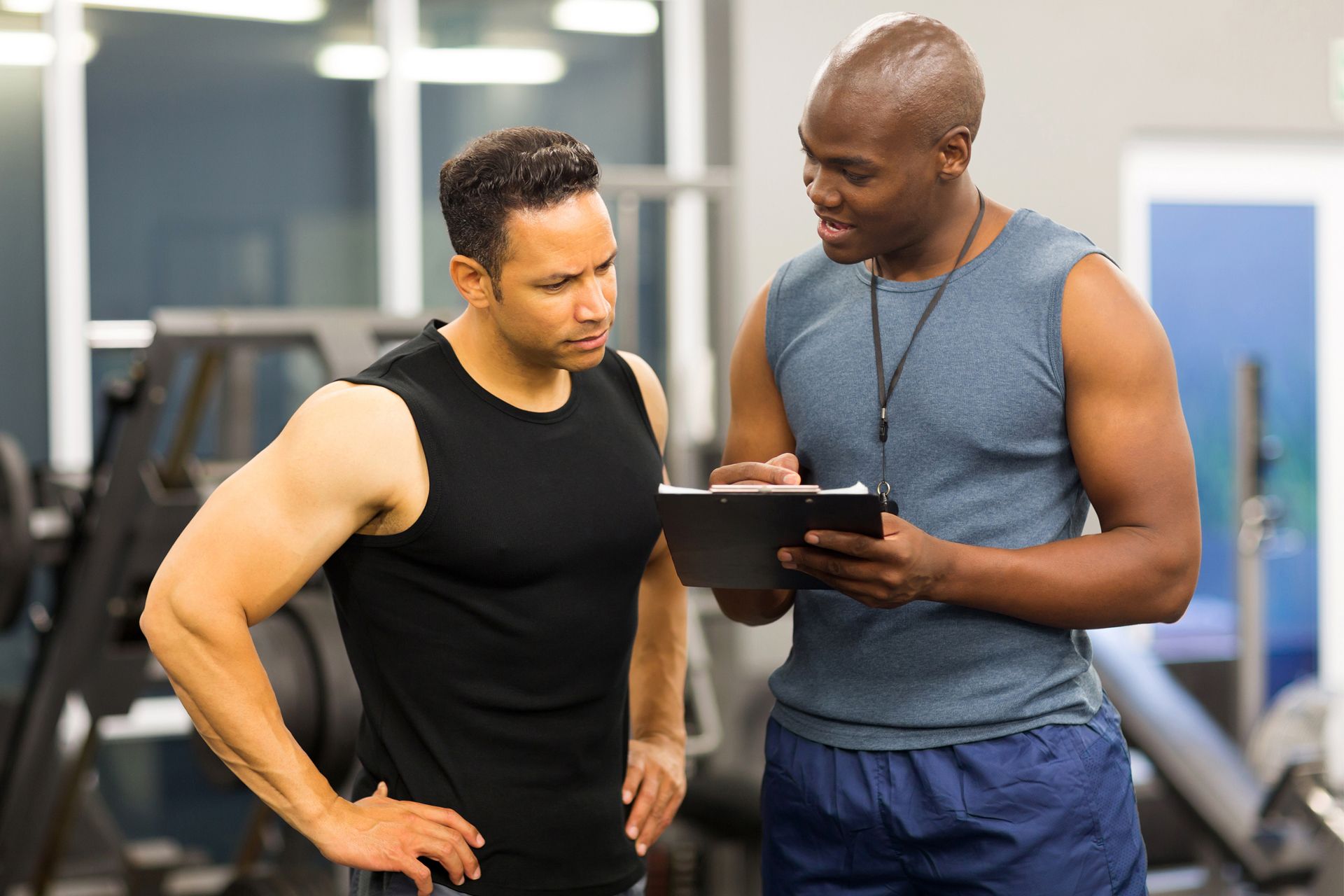 Man in black tank top consulting with a trainer in a gym, trainer holding a clipboard.