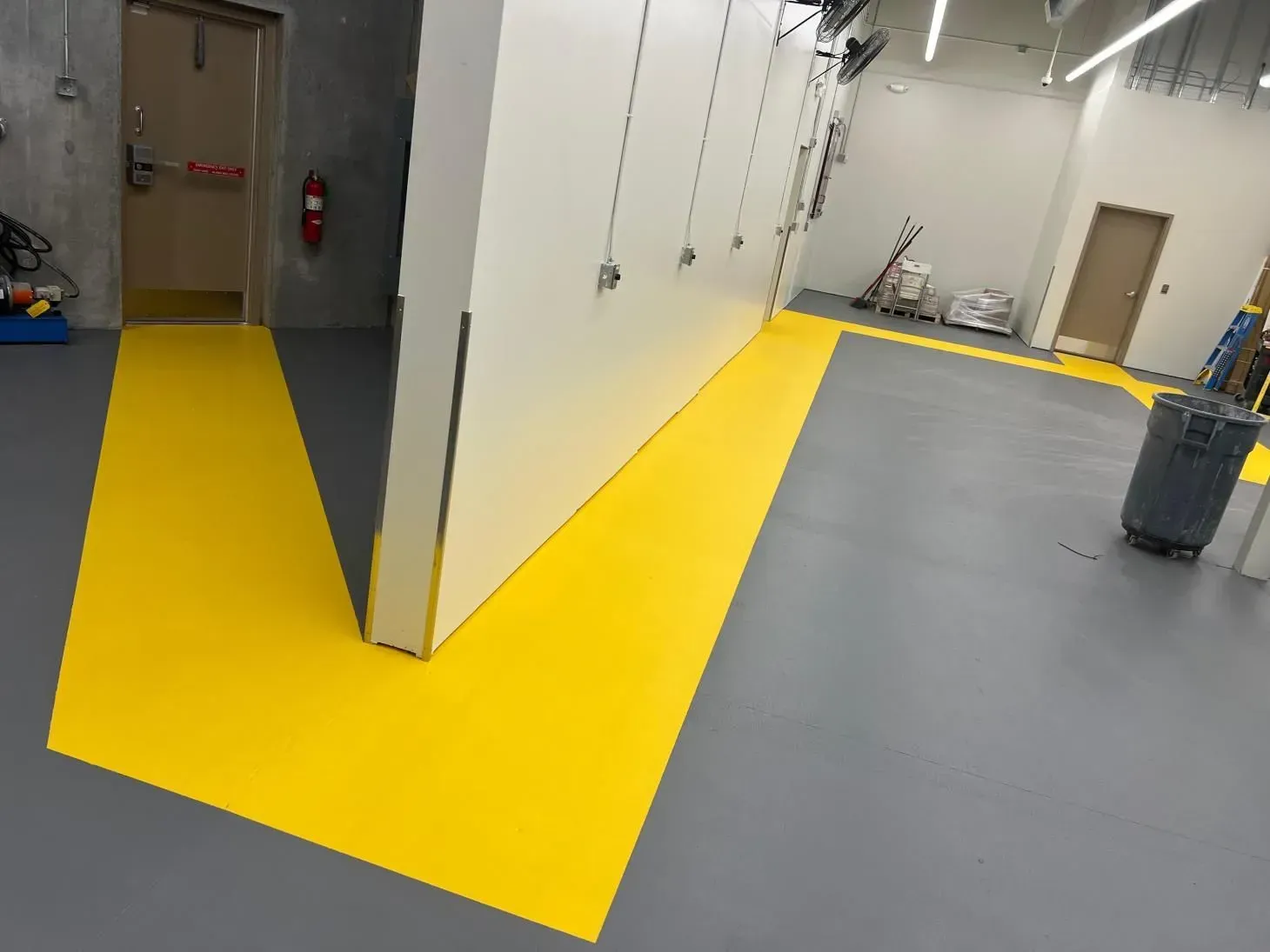 Yellow pathway guides through a gray-floored room with a white wall, leading to a door.