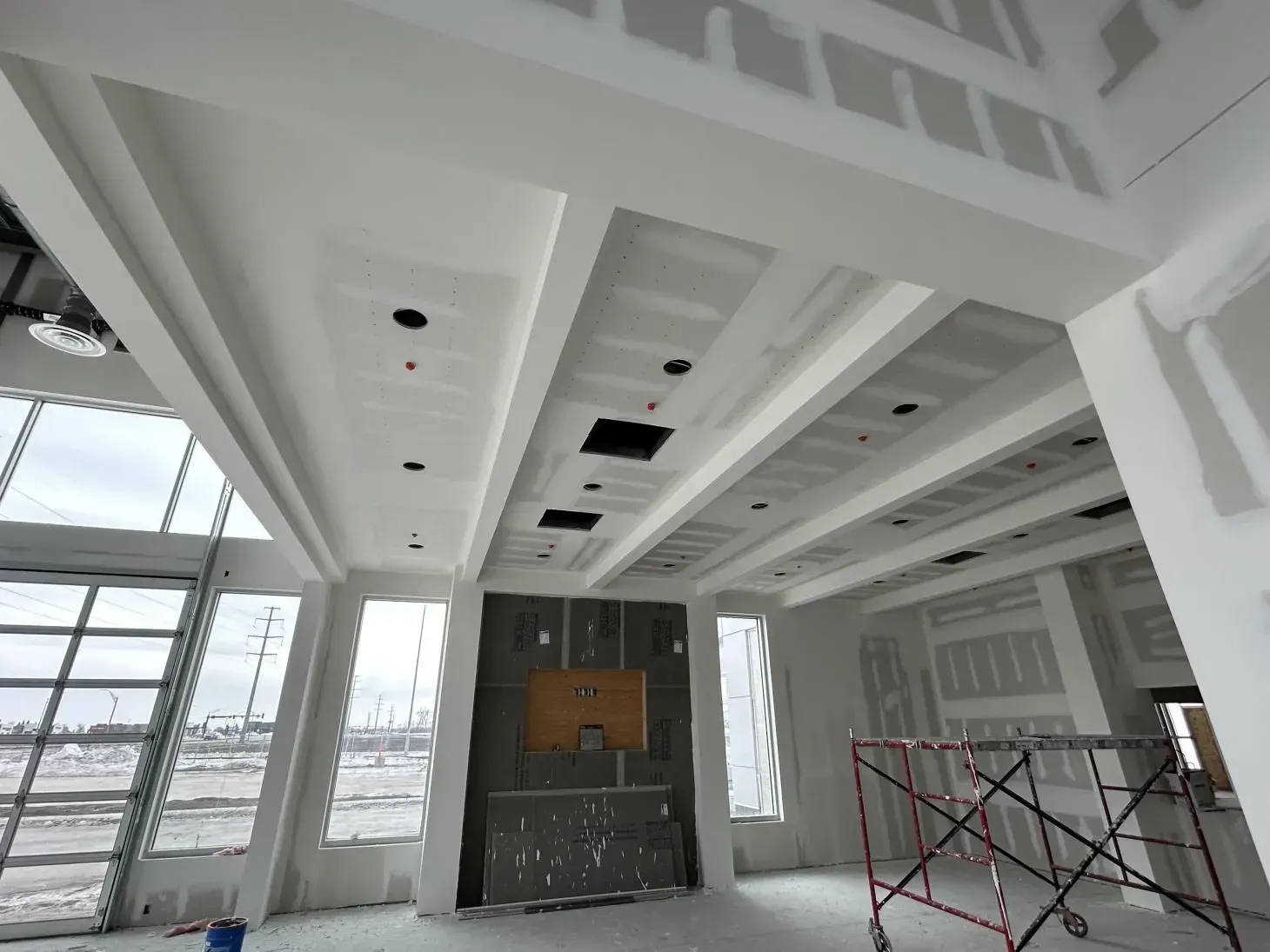 Interior under construction: drywall, ceiling beams, recessed lighting, windows, scaffolding, unfinished fireplace.