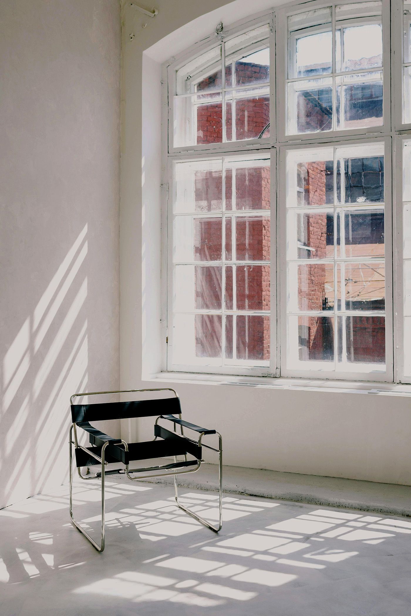 Minimalist room with a black and white chair in front of a window; sunlight casts shadows on the floor and wall.