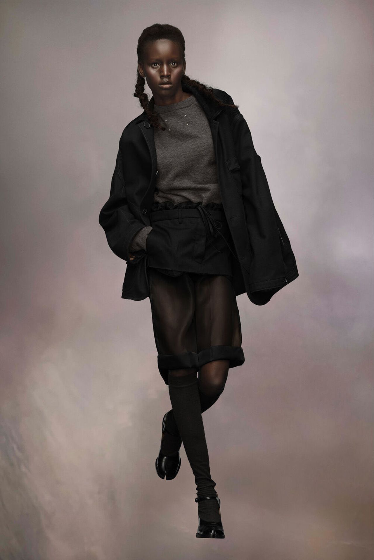 Model in dark outfit: coat, sweater, sheer skirt, knee-high boots, against a muted background.