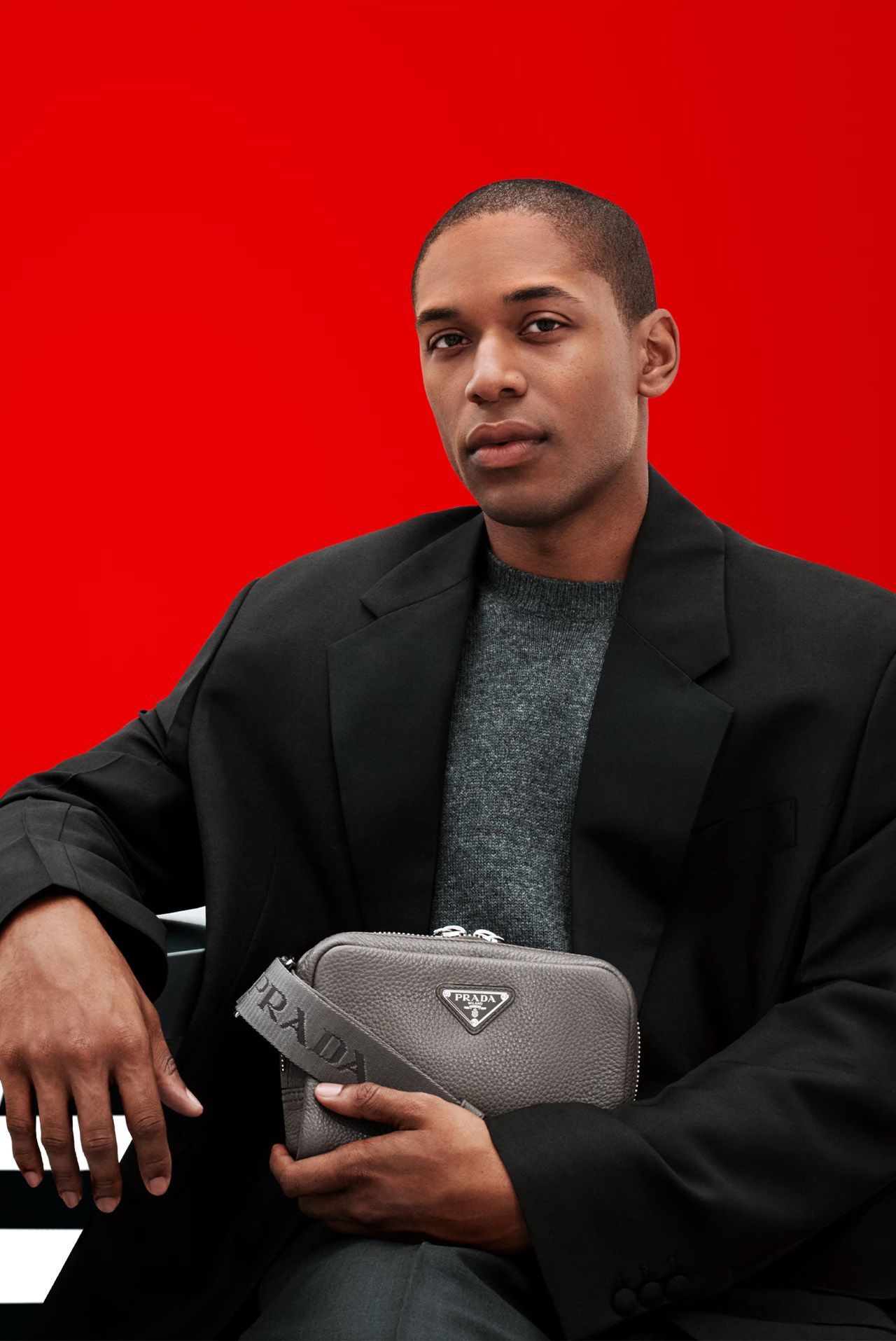 Man holding a gray bag with a Prada logo, wearing a black blazer and gray sweater, against a red backdrop.
