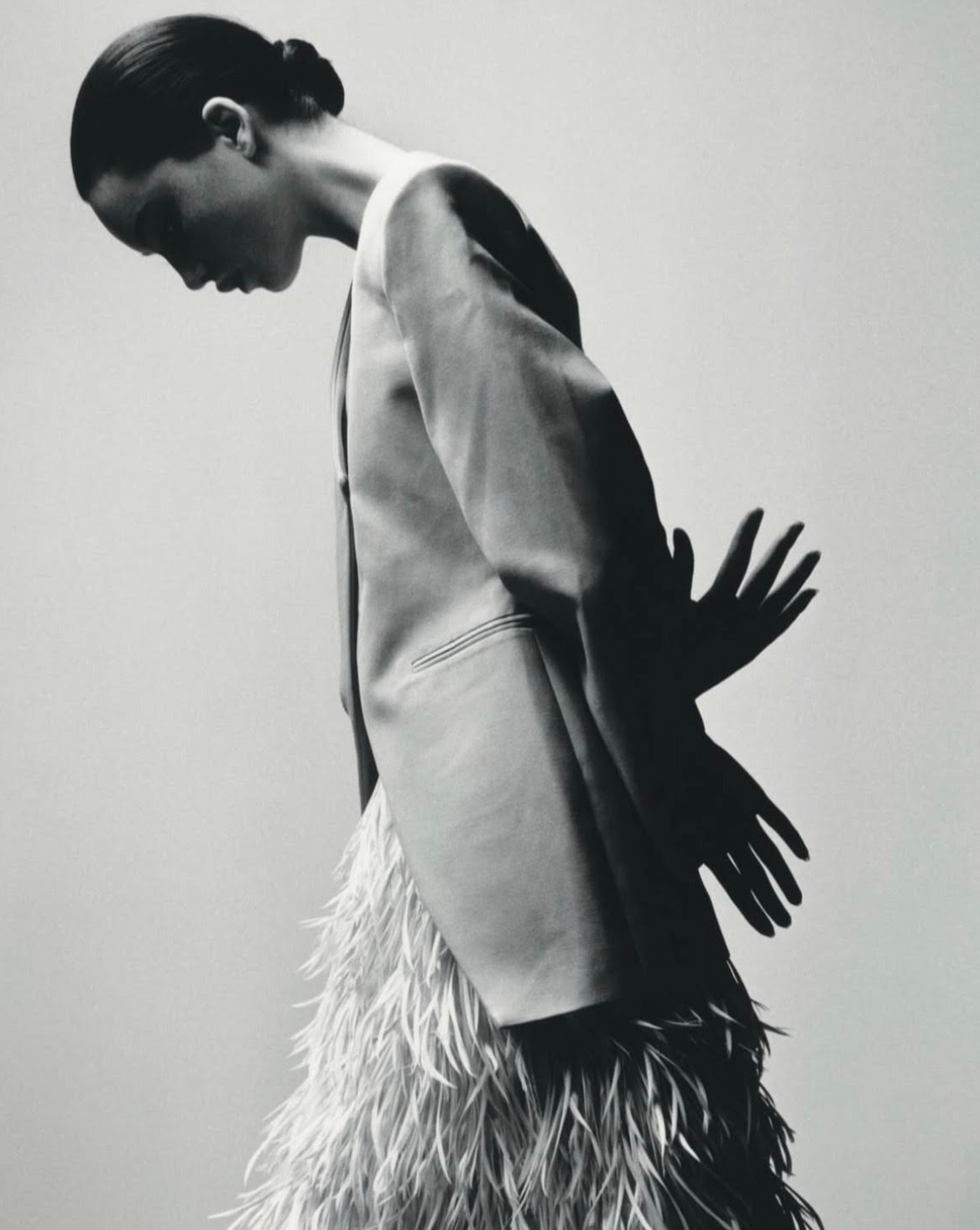 Woman in tailored jacket and feathered skirt, hands behind back, head bowed. Black and white.