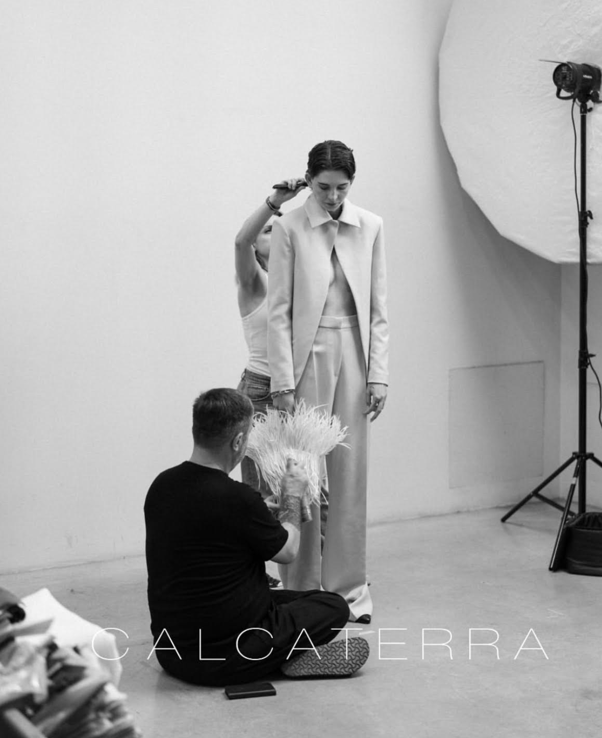 Model in beige suit, being adjusted by stylist, photographed in studio setting.