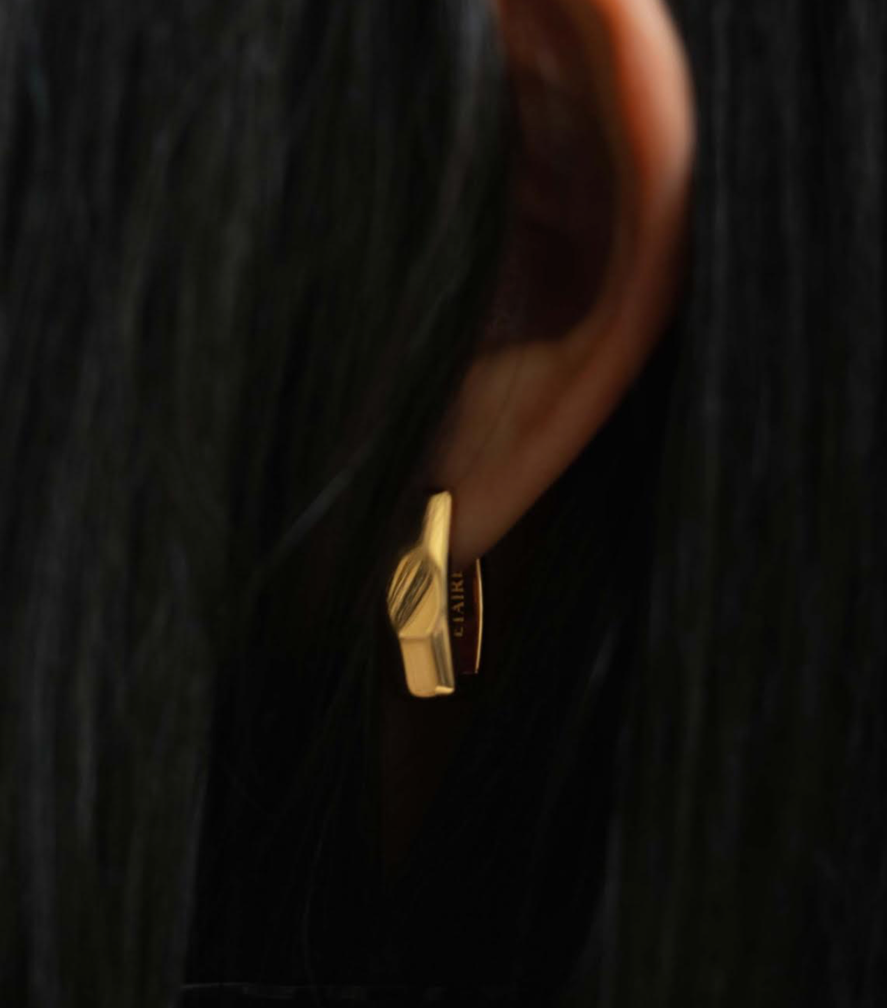 Close-up of a gold earring on an ear. The earring is a small, geometric shape with a leaf design.