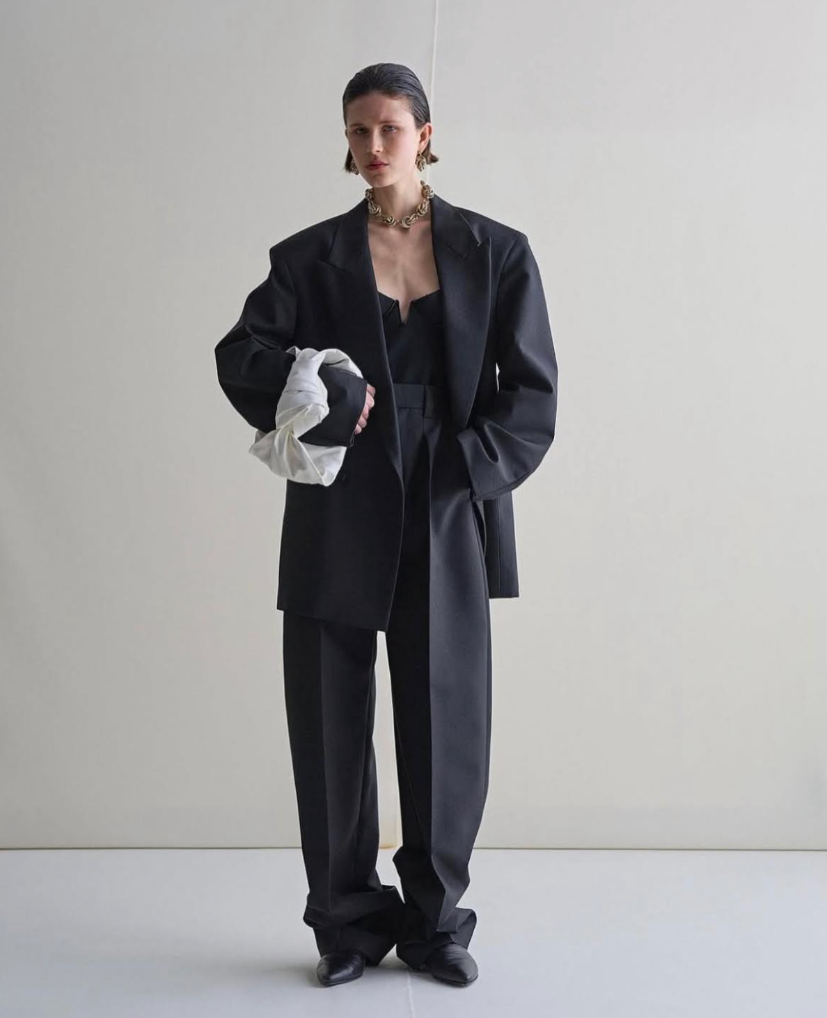 Woman in black blazer, suit pants, and corset top, holding white ruffled clutch, poses against a plain backdrop.