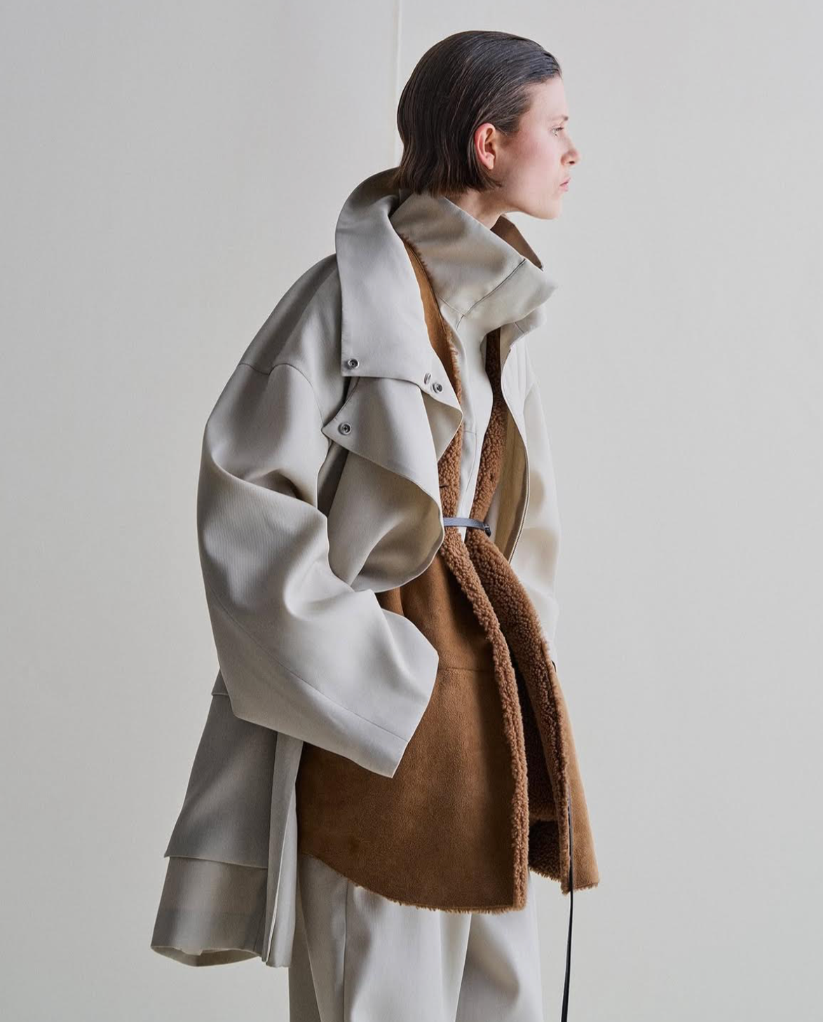 Woman wearing layered beige coat and brown fluffy bag, standing, side view.