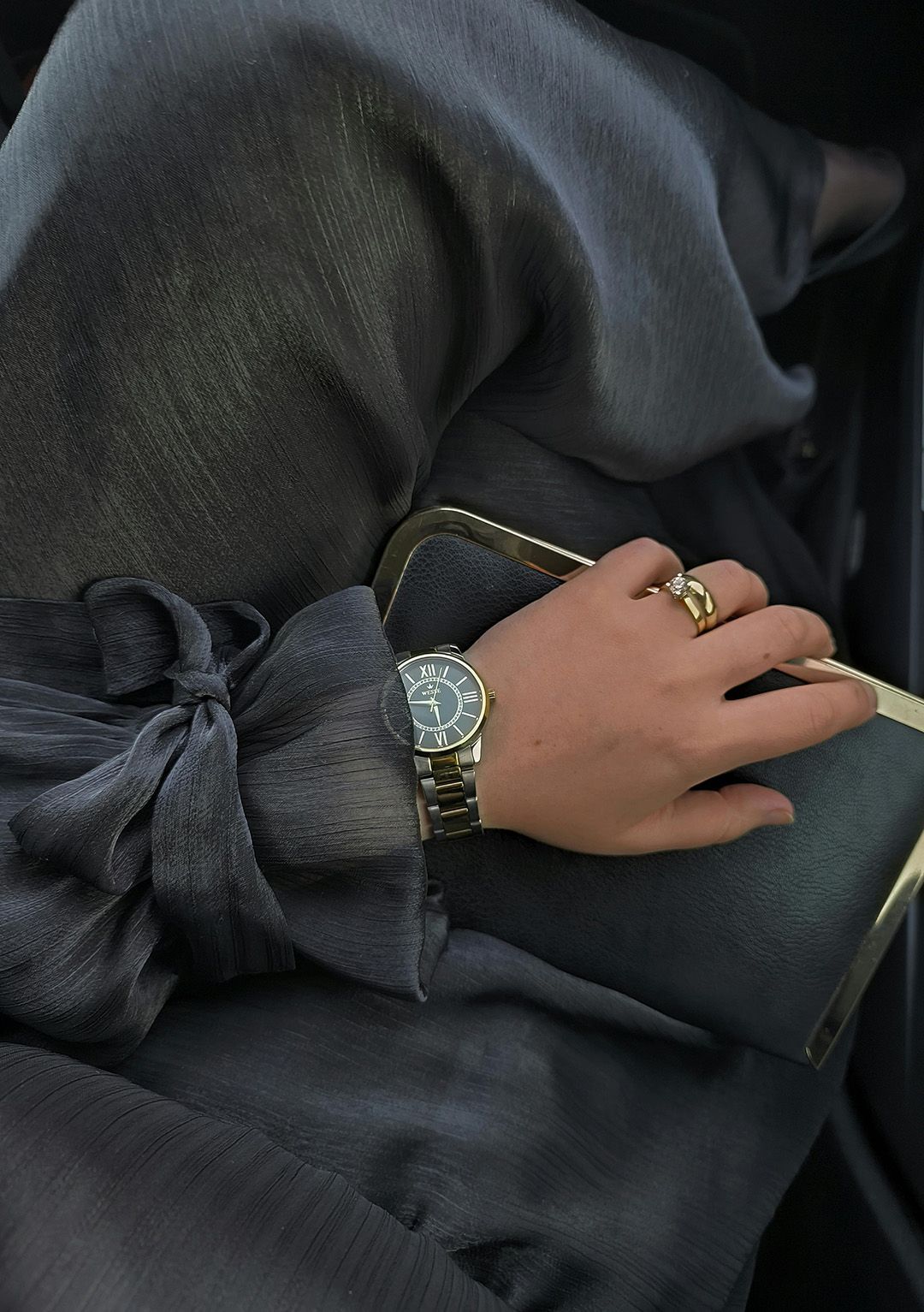 Woman's arm with a bow sleeve, watch, and rings holding a clutch, resting on dark fabric.