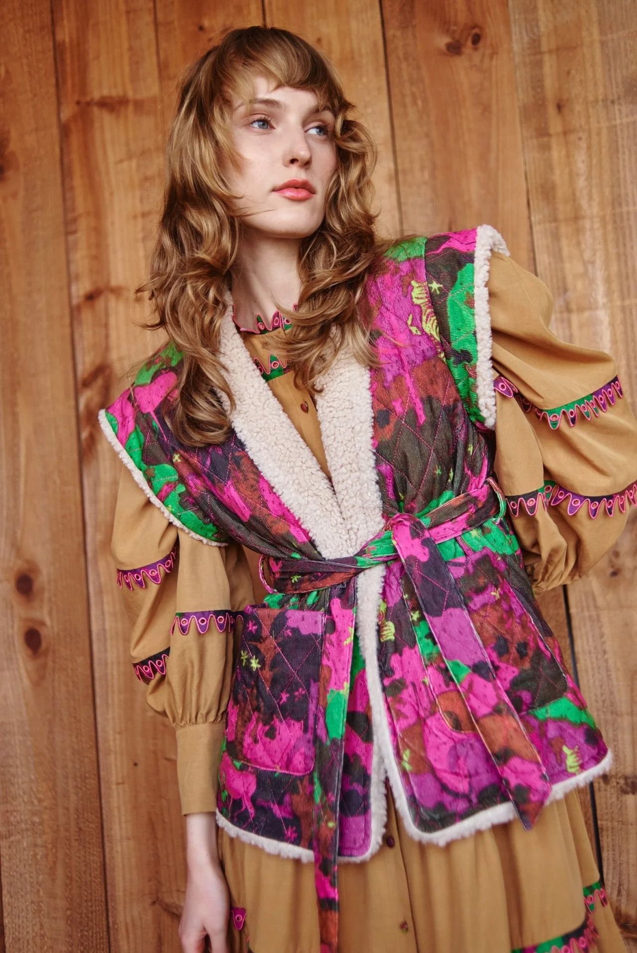 Woman in floral vest, brown dress, standing against wood paneling.