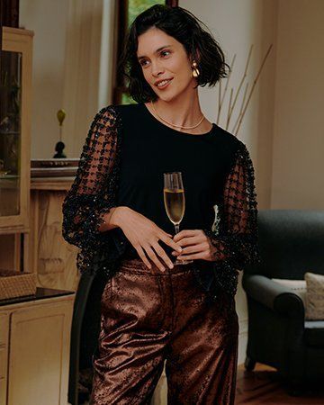 Woman in black top and bronze pants holds champagne flute, standing indoors.