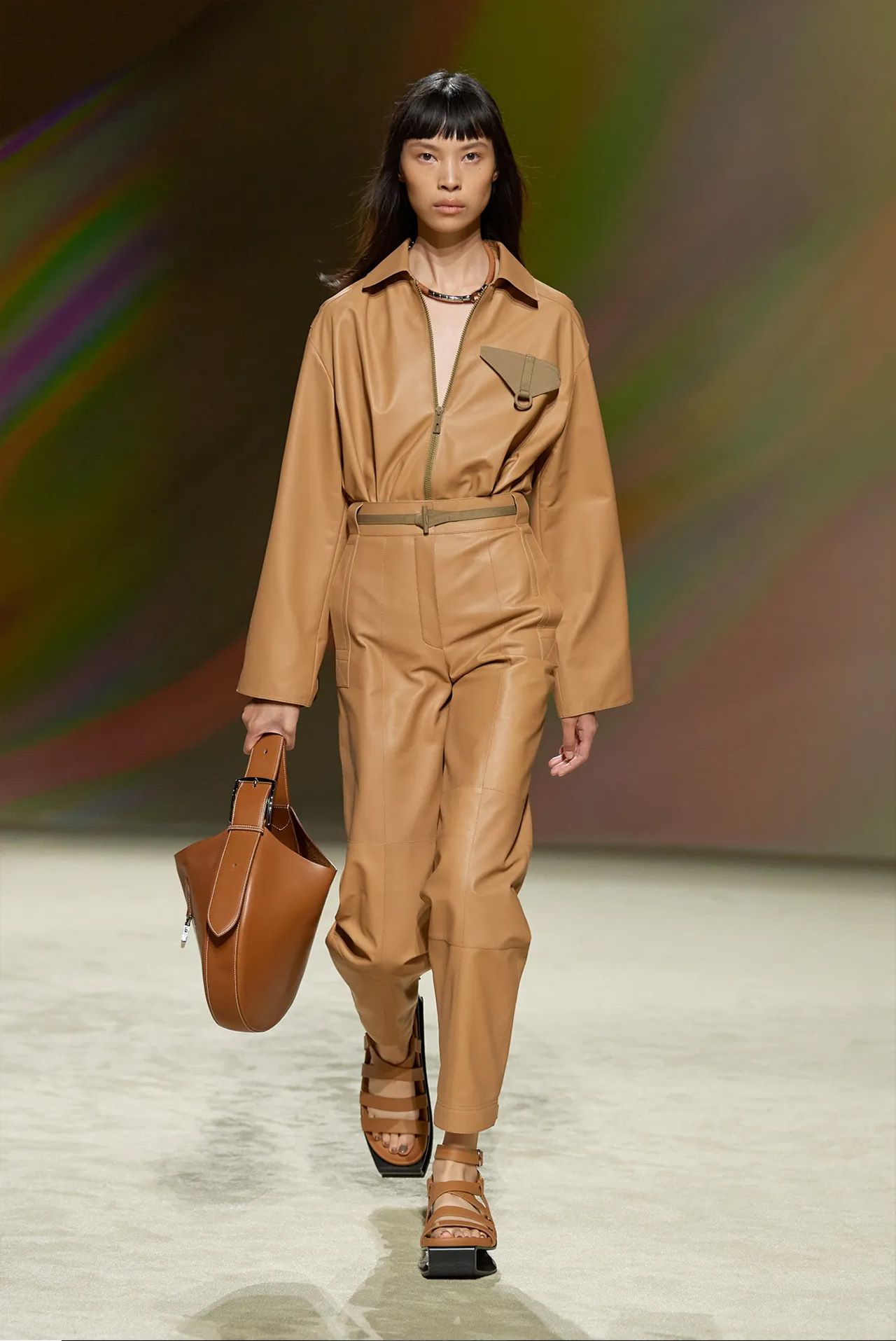Model wearing a tan leather jumpsuit and sandals, carrying a brown purse, walking a runway.
