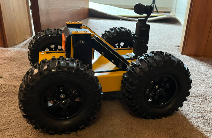 Yellow and black robot with large tires and a camera on a carpeted surface.