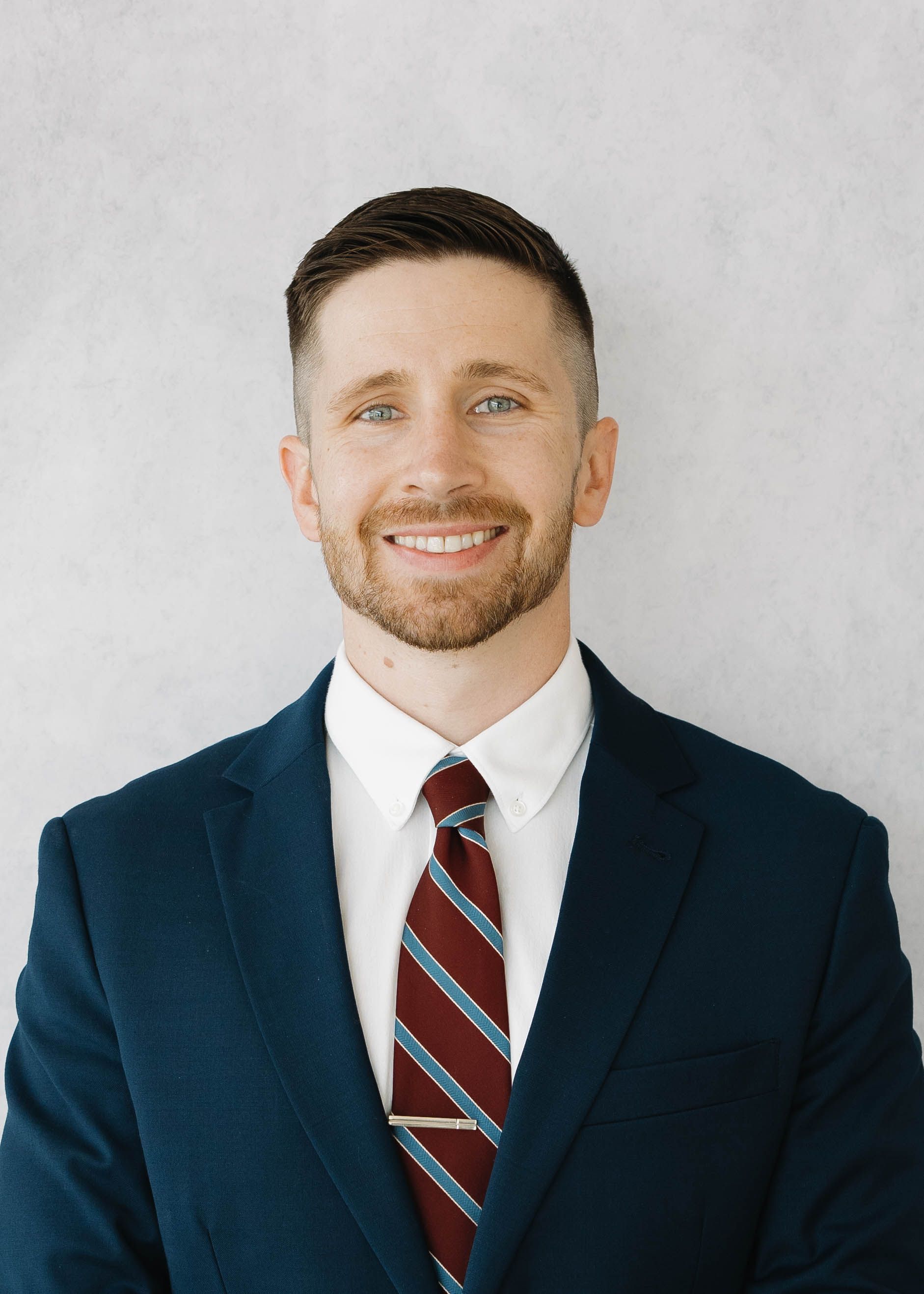 Smiling person in a navy suit, white shirt, and red striped tie against a light gray backdrop