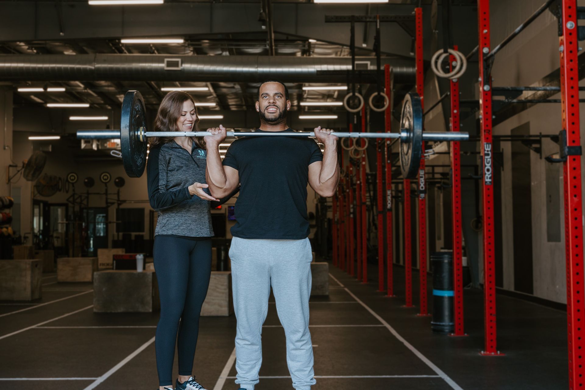 Two people holding a barbell overhead in a gym with red equipment and a dark industrial interior