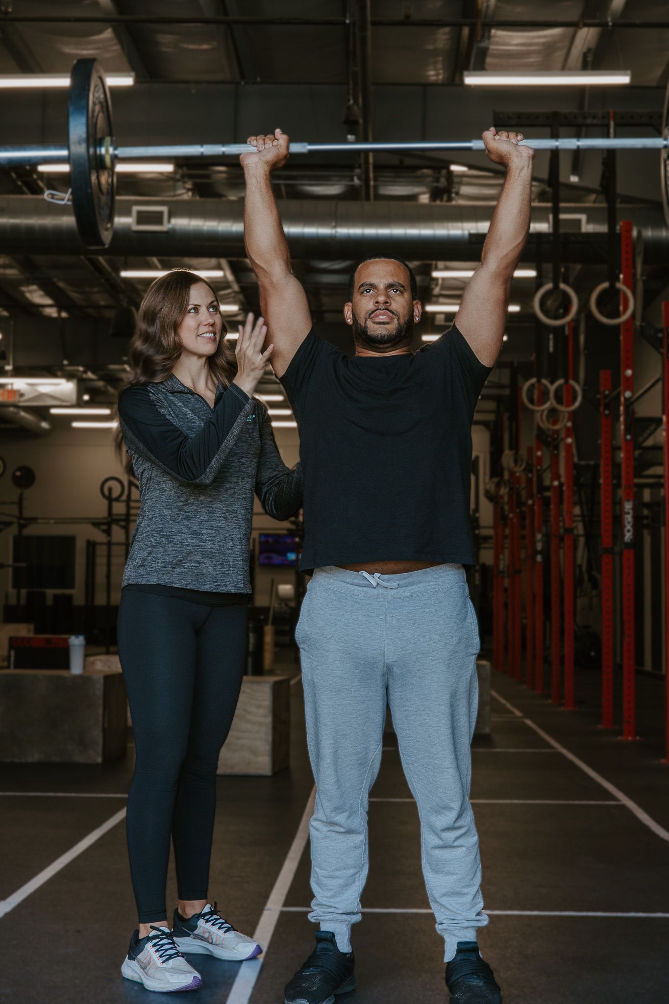 Two people standing in a gym, smiling and holding a barbell overhead