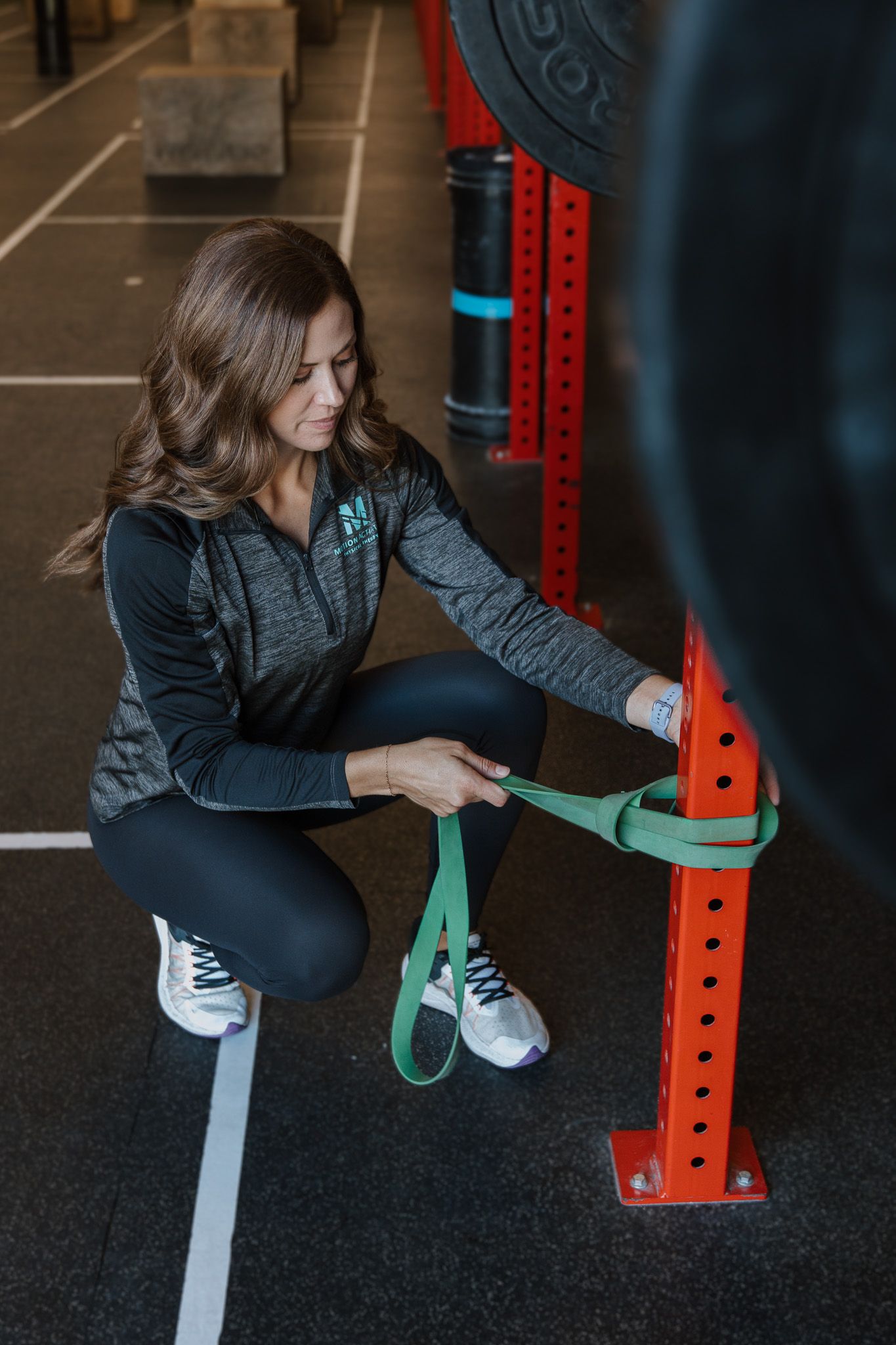 Woman kneeling beside a red gym rack, adjusting a green resistance band in a fitness gym.