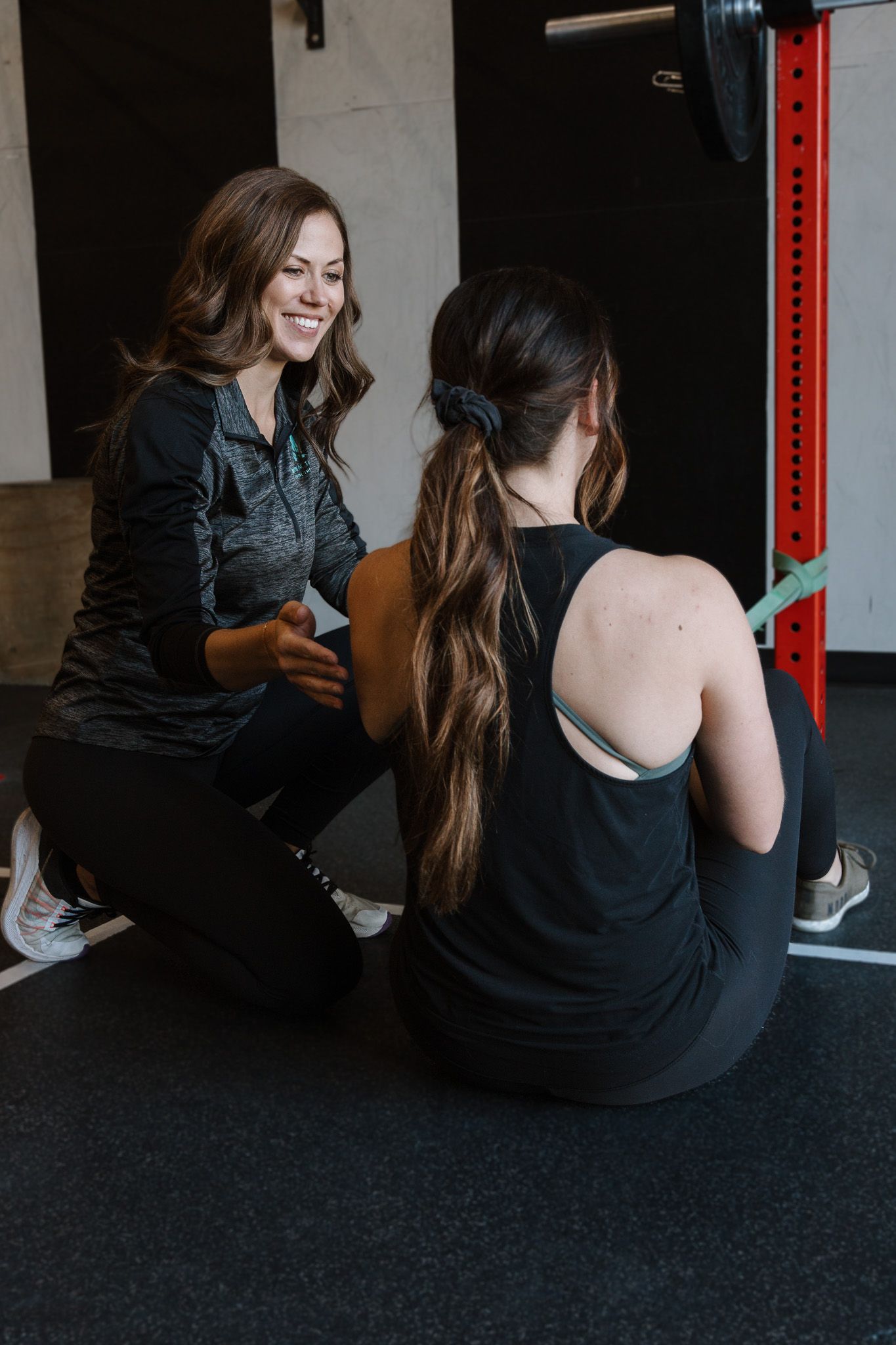 Two people sitting on a gym floor, one smiling and talking beside a red and black rack.