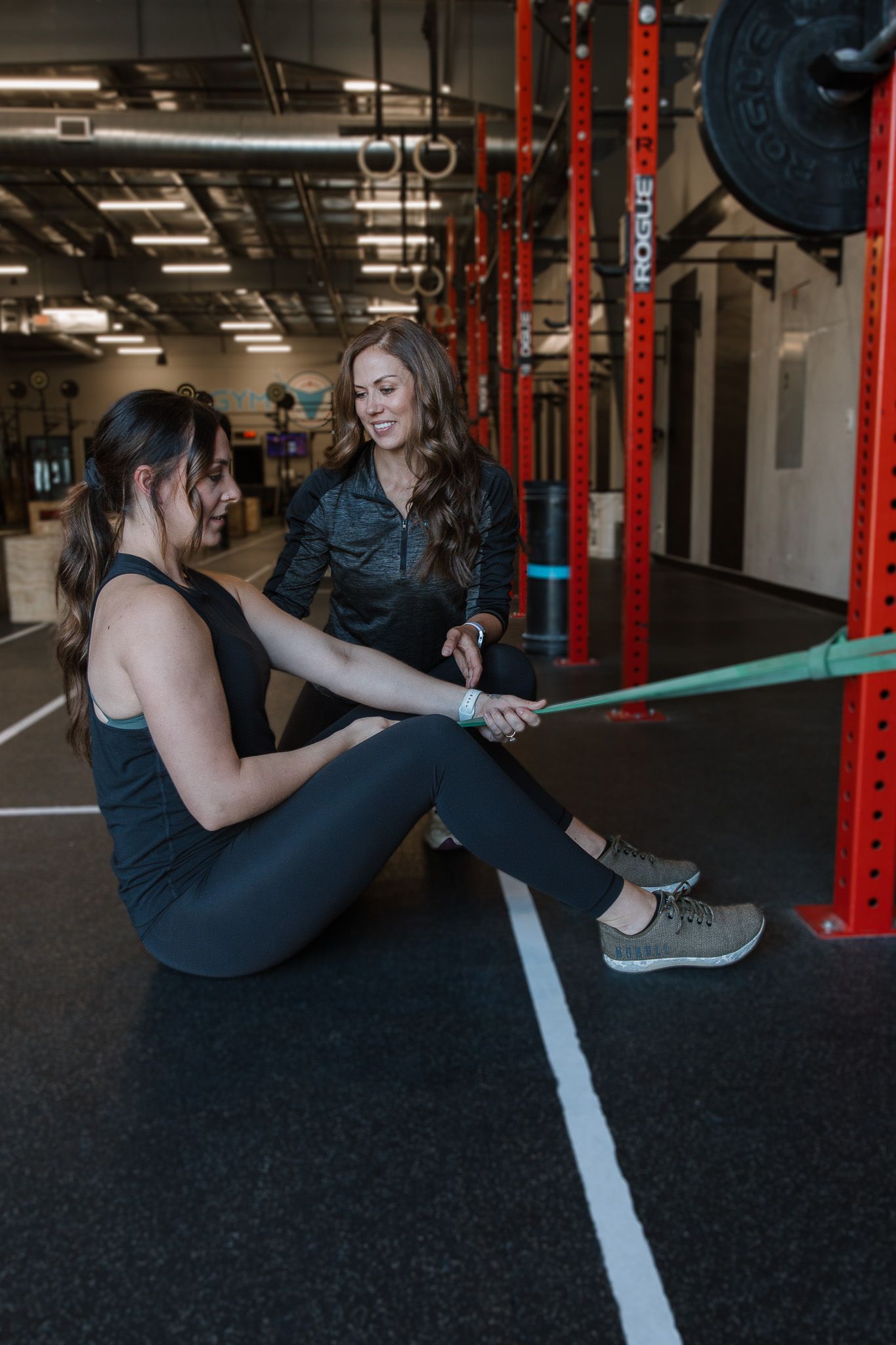 Two women exercising with a resistance band in a gym, seated on the floor near weight racks.
