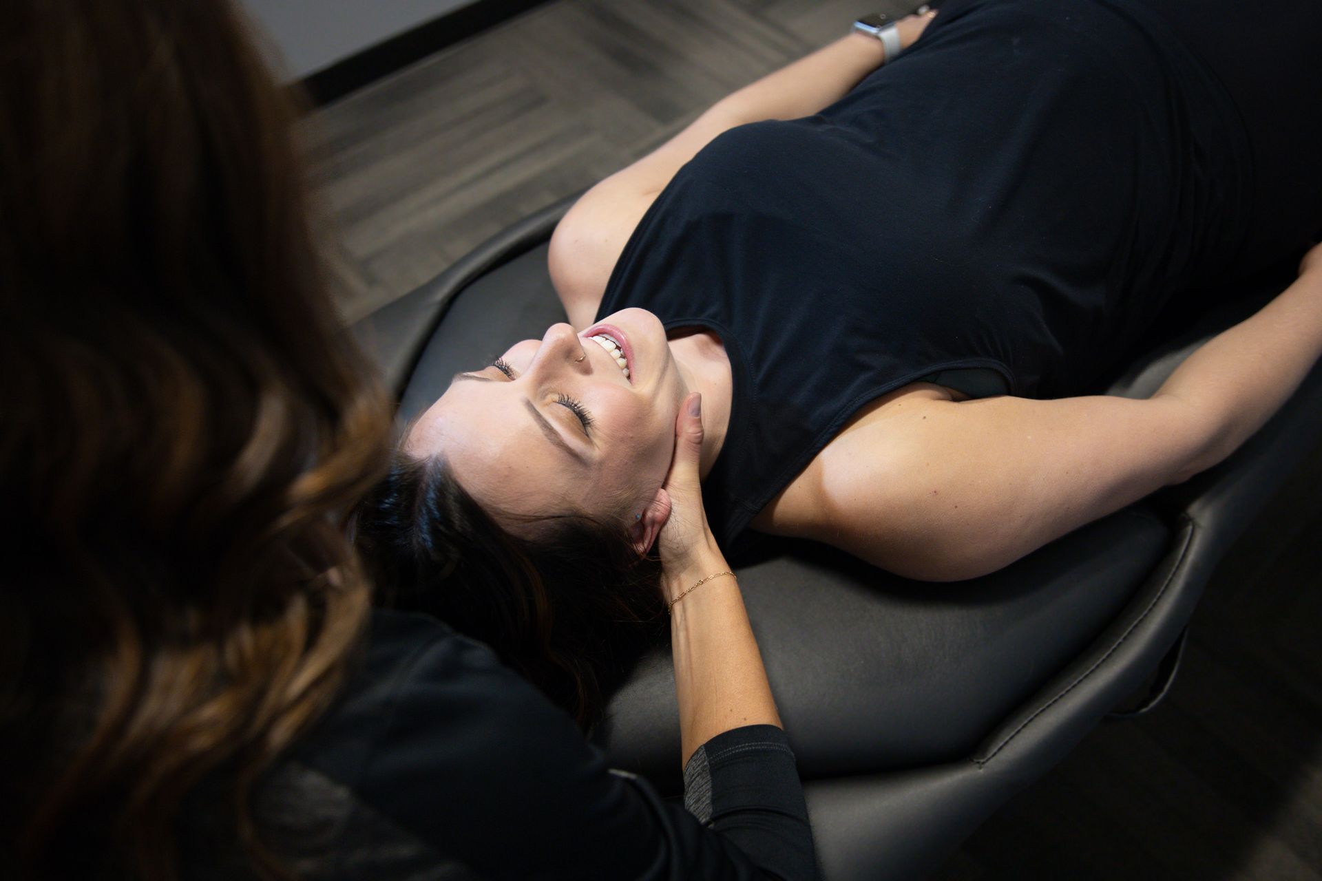 Woman lying on a black massage chair while a therapist supports her head.
