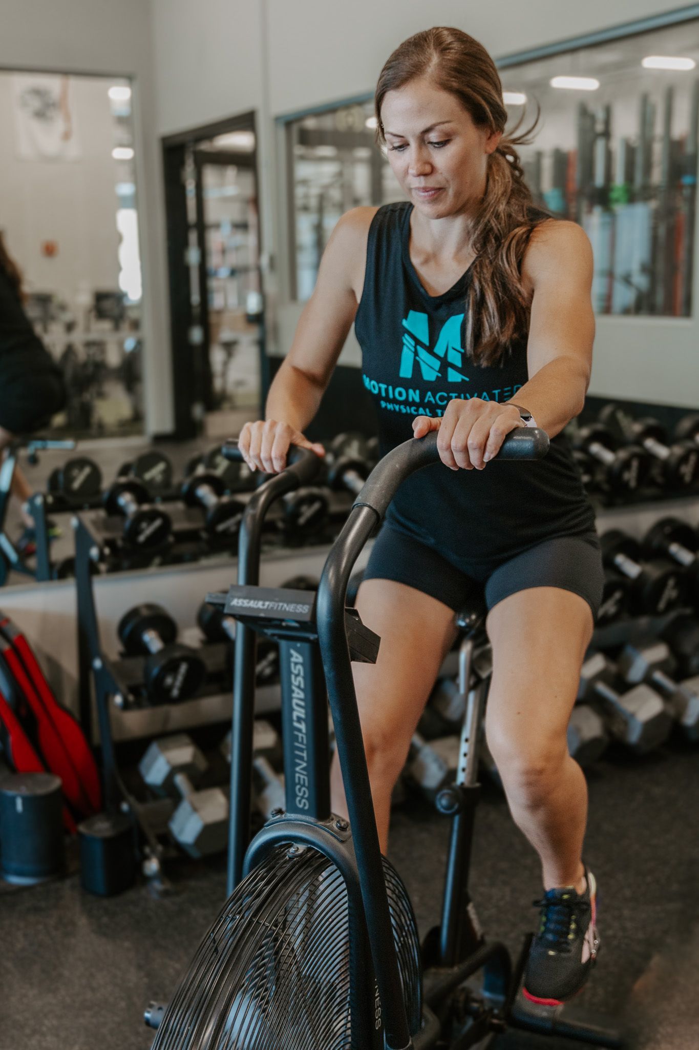 Woman exercising on an air bike in a gym, wearing a black tank top and shorts.