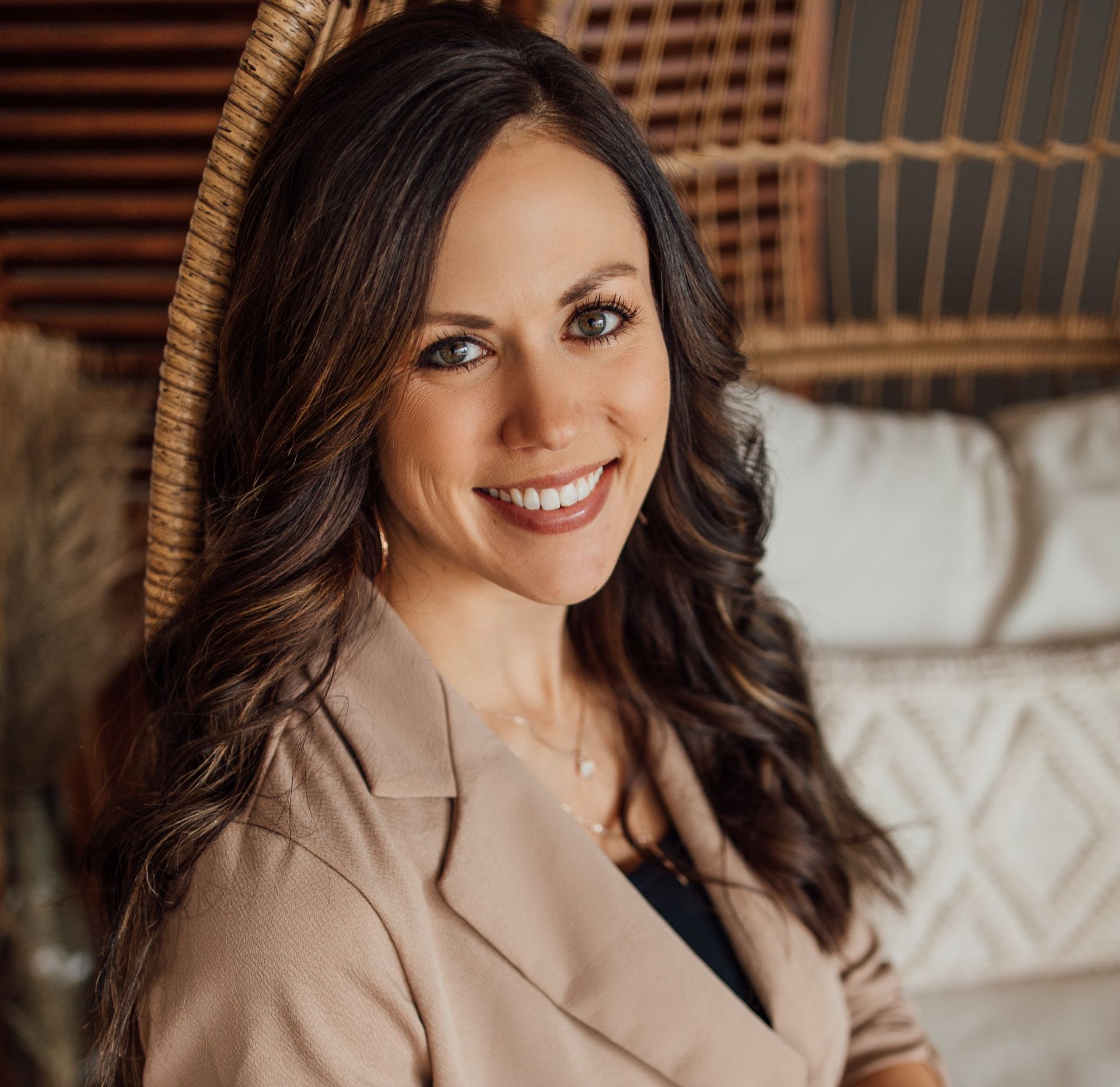 Smiling woman in beige blazer seated indoors against a wicker headboard