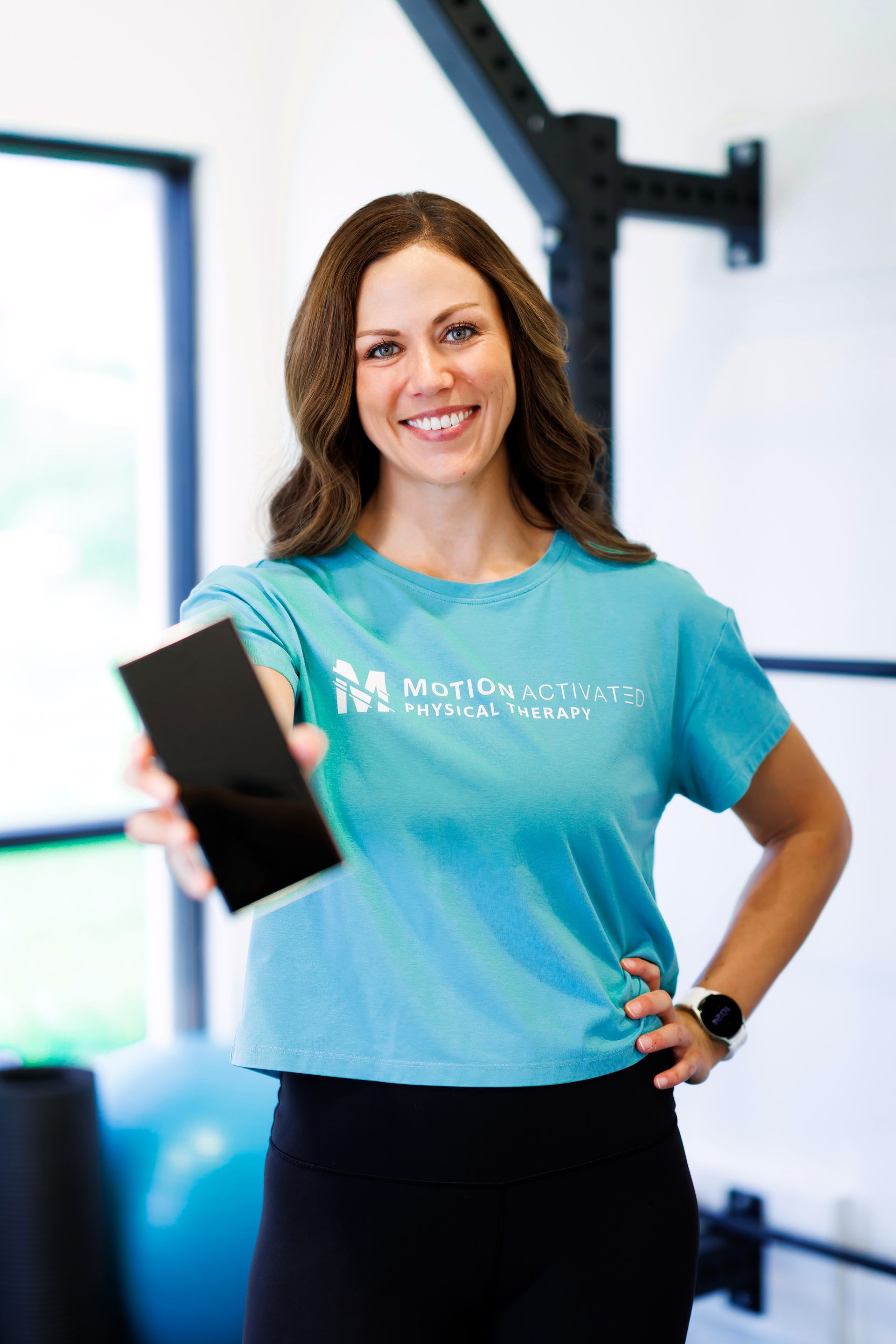 Woman in teal shirt holding a smartphone in a gym, smiling at the camera.