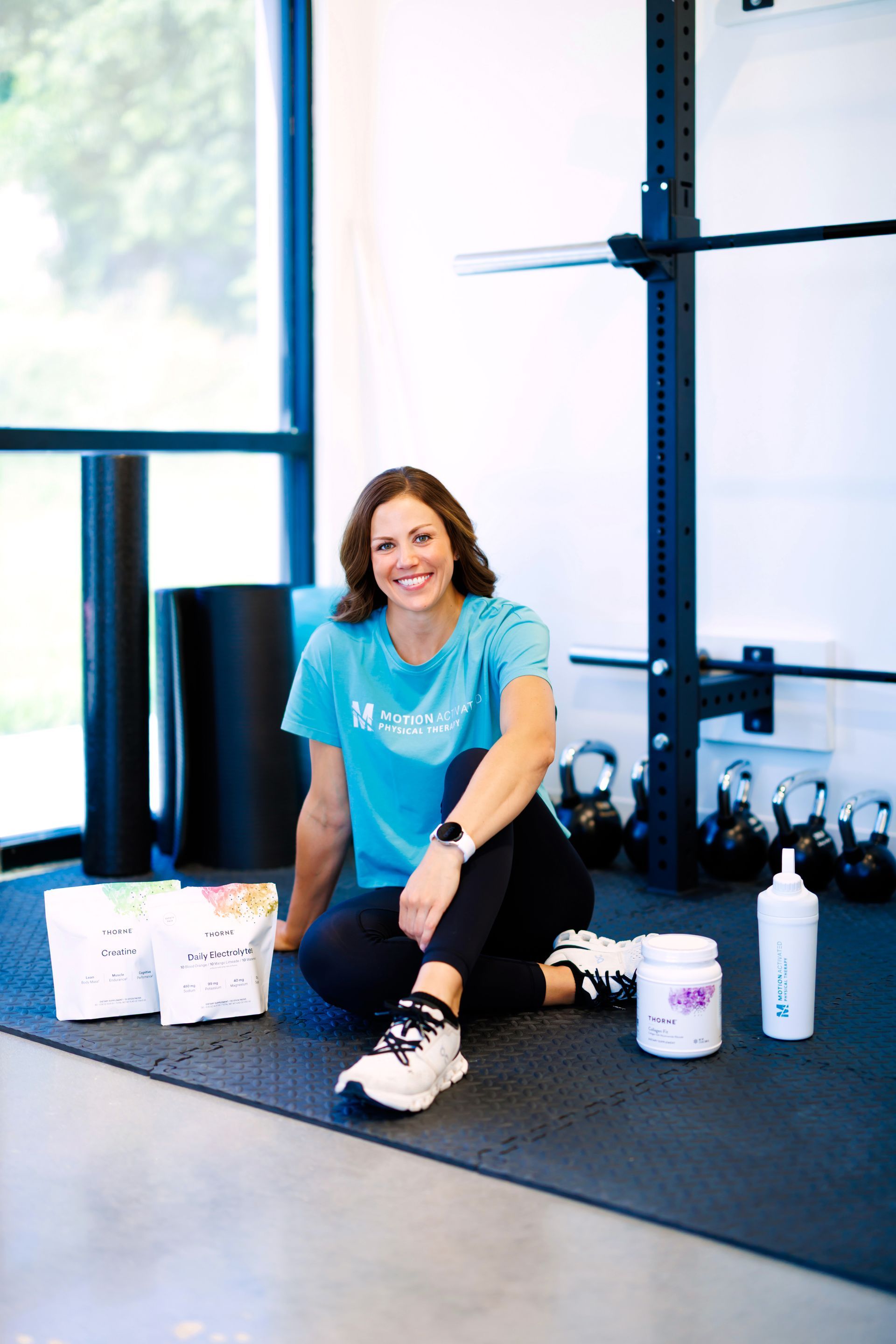 Woman sitting on a gym floor beside workout gear and a water bottle, smiling in a bright studio.