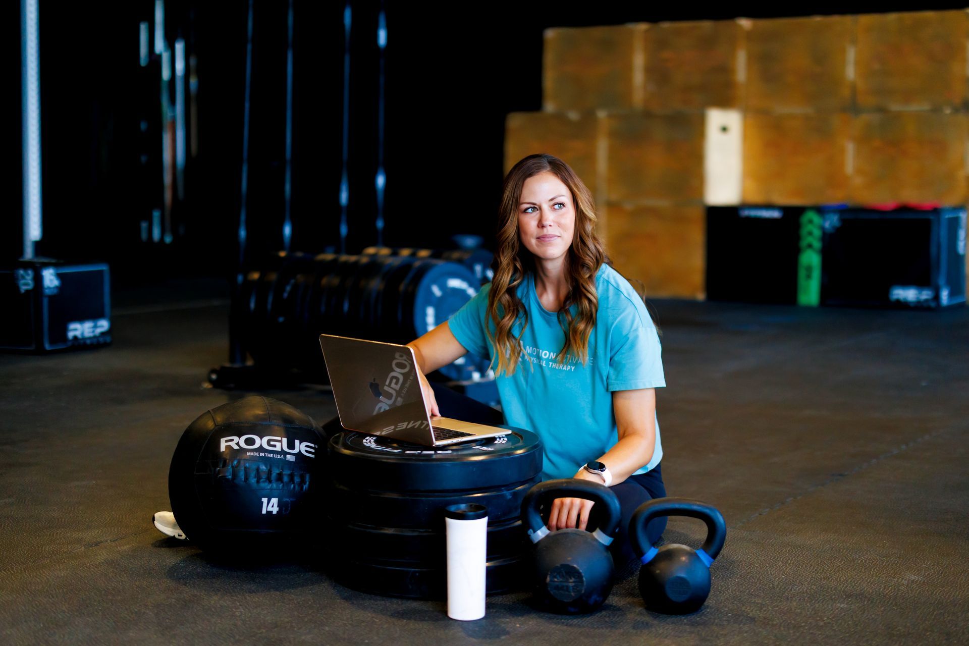 Woman sitting on a tire with a laptop and gym gear in a warehouse-like gym setting