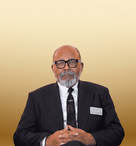 Man in a black suit, tie, and glasses stands against a gold gradient background.