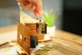 Hand placing key in a small brown wallet beside scattered coins on a wooden table