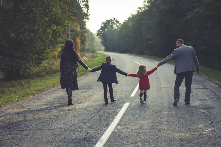 Family holding hands, walking away down a tree-lined road on a cloudy day.