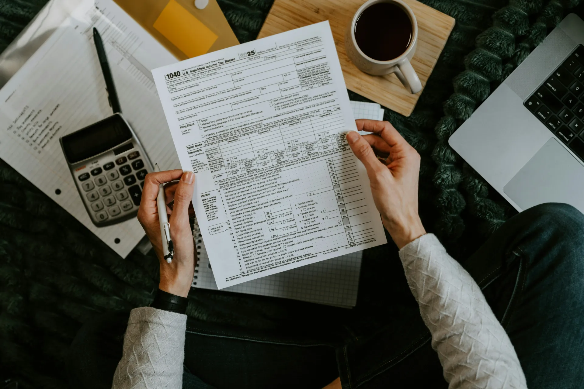 Person reviewing paperwork at a desk with calculator, coffee, laptop, and notes