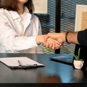 Handshake across a desk in an office, with a clipboard, laptop, and small potted plant.