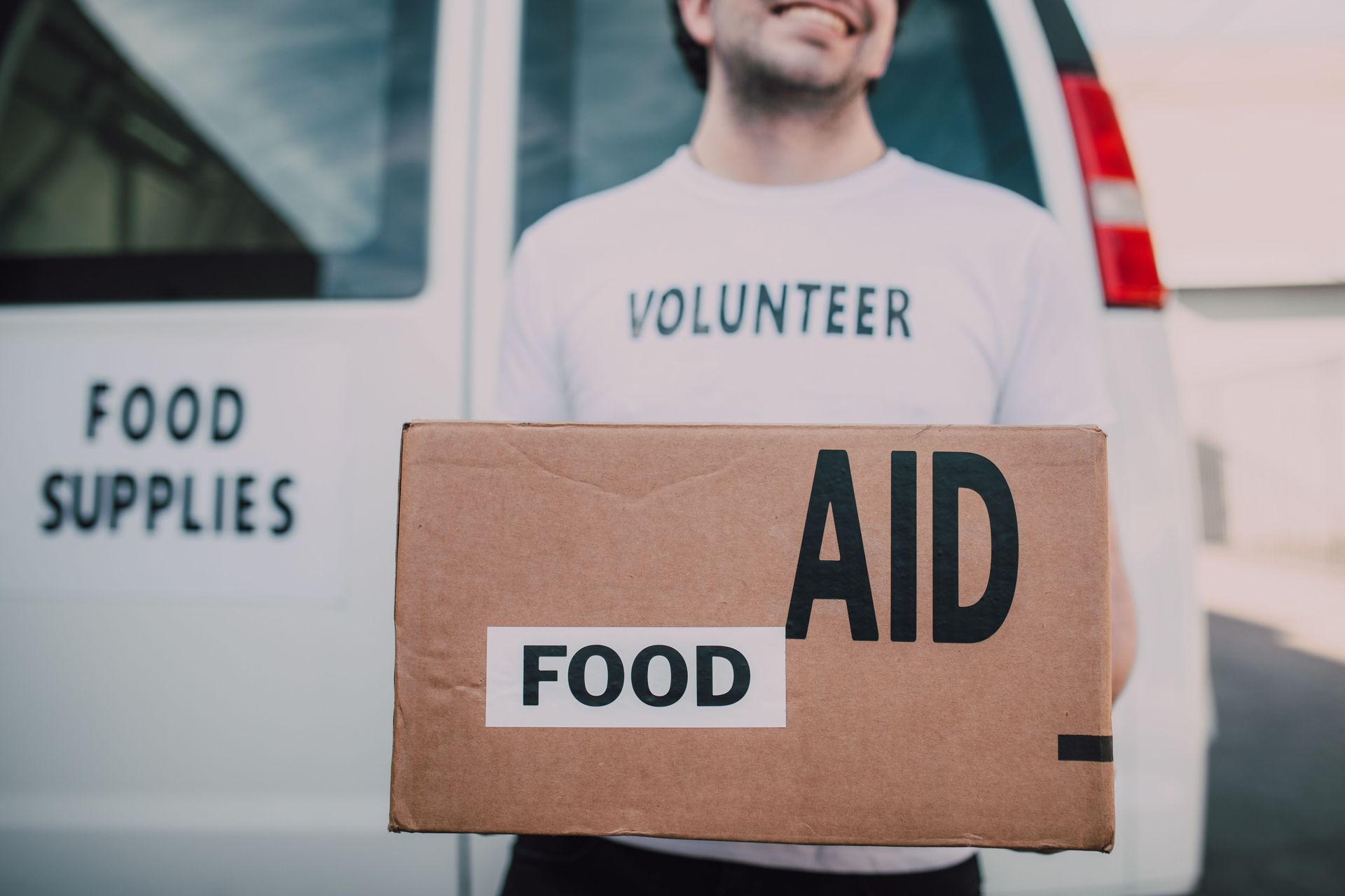 Volunteer holding cardboard box labeled “AID” and “FOOD” beside a food supplies van.