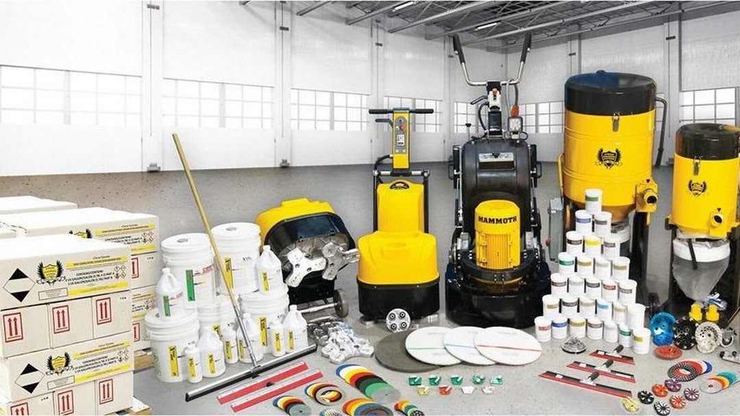 Assorted cleaning equipment and supplies arranged in a bright warehouse, including yellow floor scrubbers and buckets.